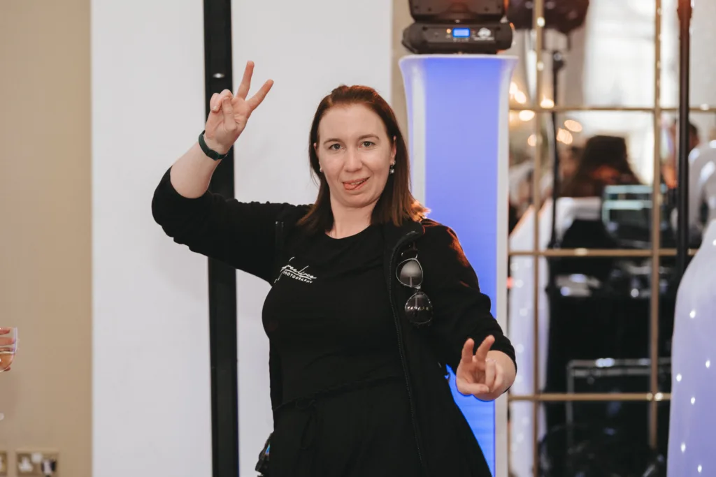 A woman in a black outfit stands indoors, making peace signs with both hands and playfully sticking out her tongue. She wears a smartwatch and sunglasses on her neckline. Behind her are mirrored panels, a blue-lit column, and reflections of other people and lights. © Aimee Lince Photography - Wedding photographer in Lincolnshire, Yorkshire & Nottinghamshire