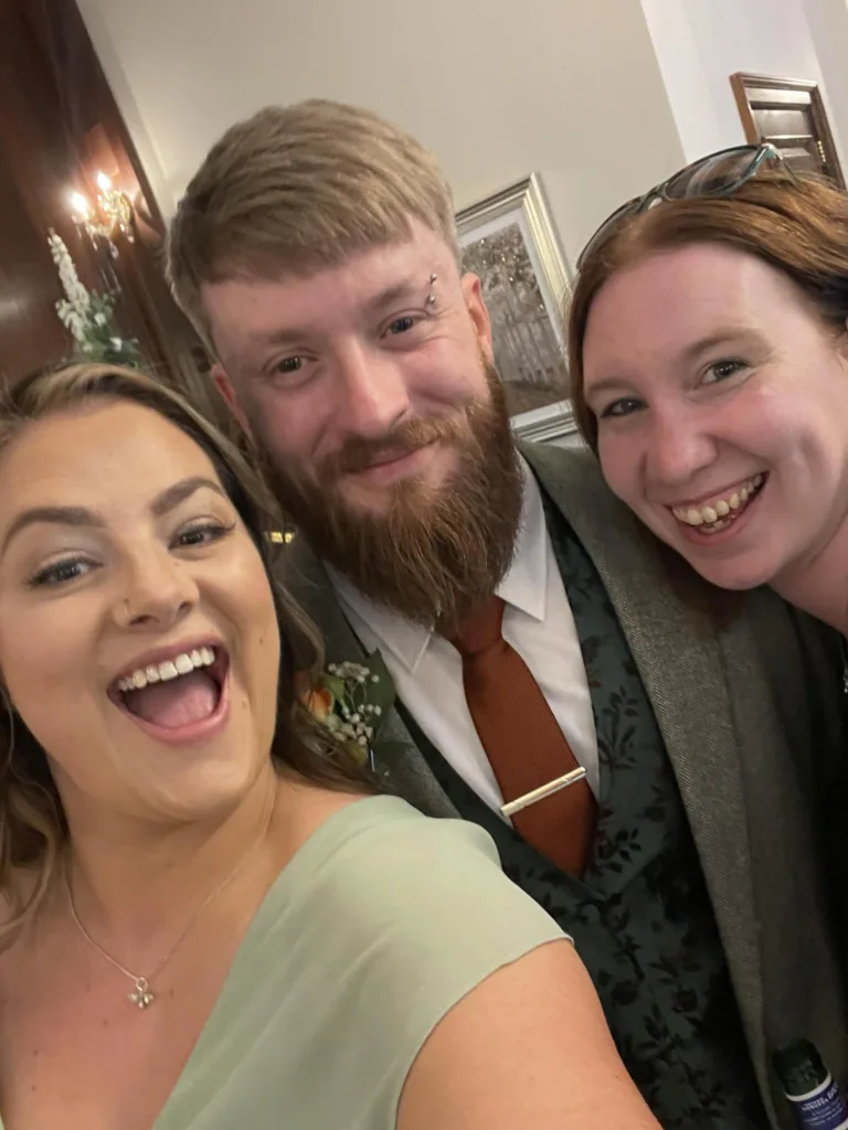 Three adults smile closely together for a selfie. The man in the middle has a beard, a reddish-brown tie, and a boutonniere. The women on either side have long hair; one wears light green, the other black. They appear happy and dressed for a formal occasion. © Aimee Lince Photography - Wedding photographer in Lincolnshire, Yorkshire & Nottinghamshire
