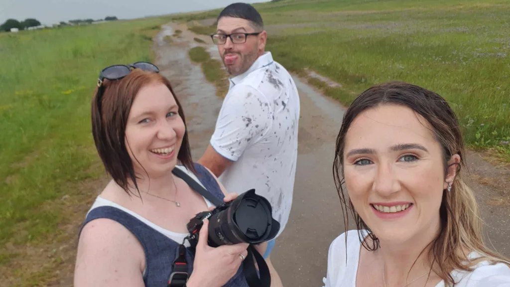 Three people stand on a wet country path. Two women in front smile at the camera; one holds a camera. A man behind them, with glasses and a beard, playfully sticks out his tongue. All wear casual clothes and appear damp from rain. Green fields stretch behind them. © Aimee Lince Photography - Wedding photographer in Lincolnshire, Yorkshire & Nottinghamshire