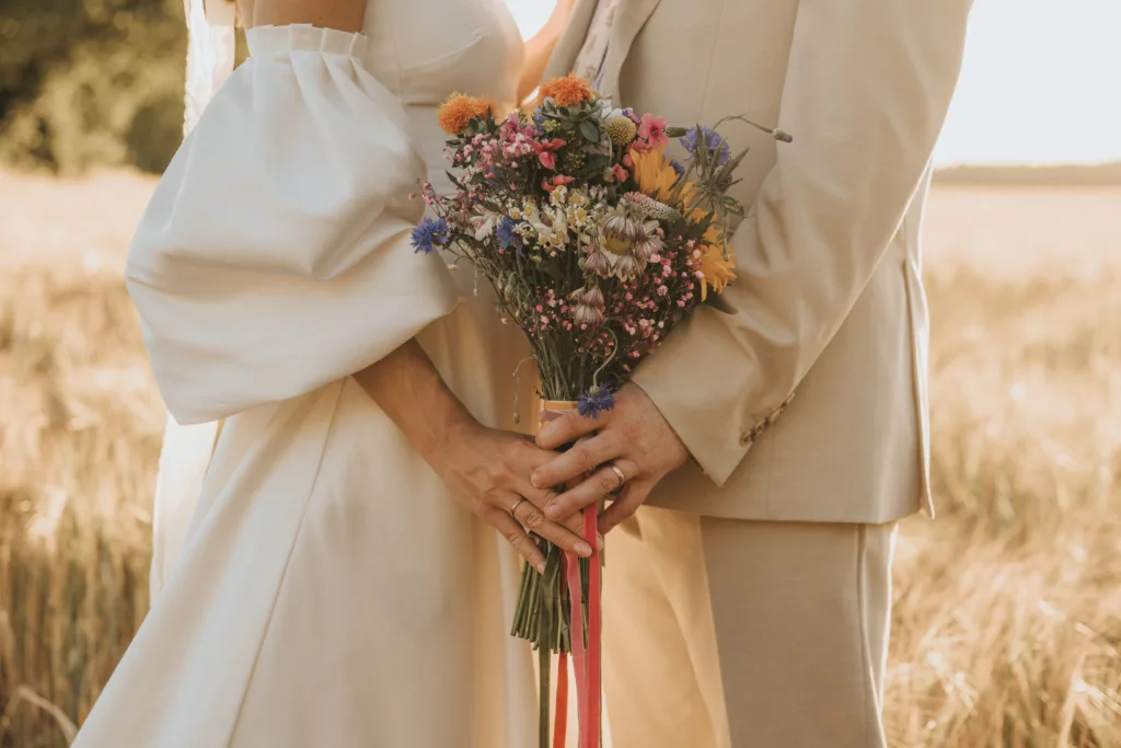 A couple in cream-colored wedding attire stands in a sunlit field, holding a vibrant bouquet of wildflowers and ribbons. A wedding photographer captures their hands, rings, and flowers, highlighting love and intimacy in a warm, romantic atmosphere. © Aimee Lince Photography - Wedding photographer in Lincolnshire, Yorkshire & Nottinghamshire. Wedding Photography Costs explained by Aimee Lince.