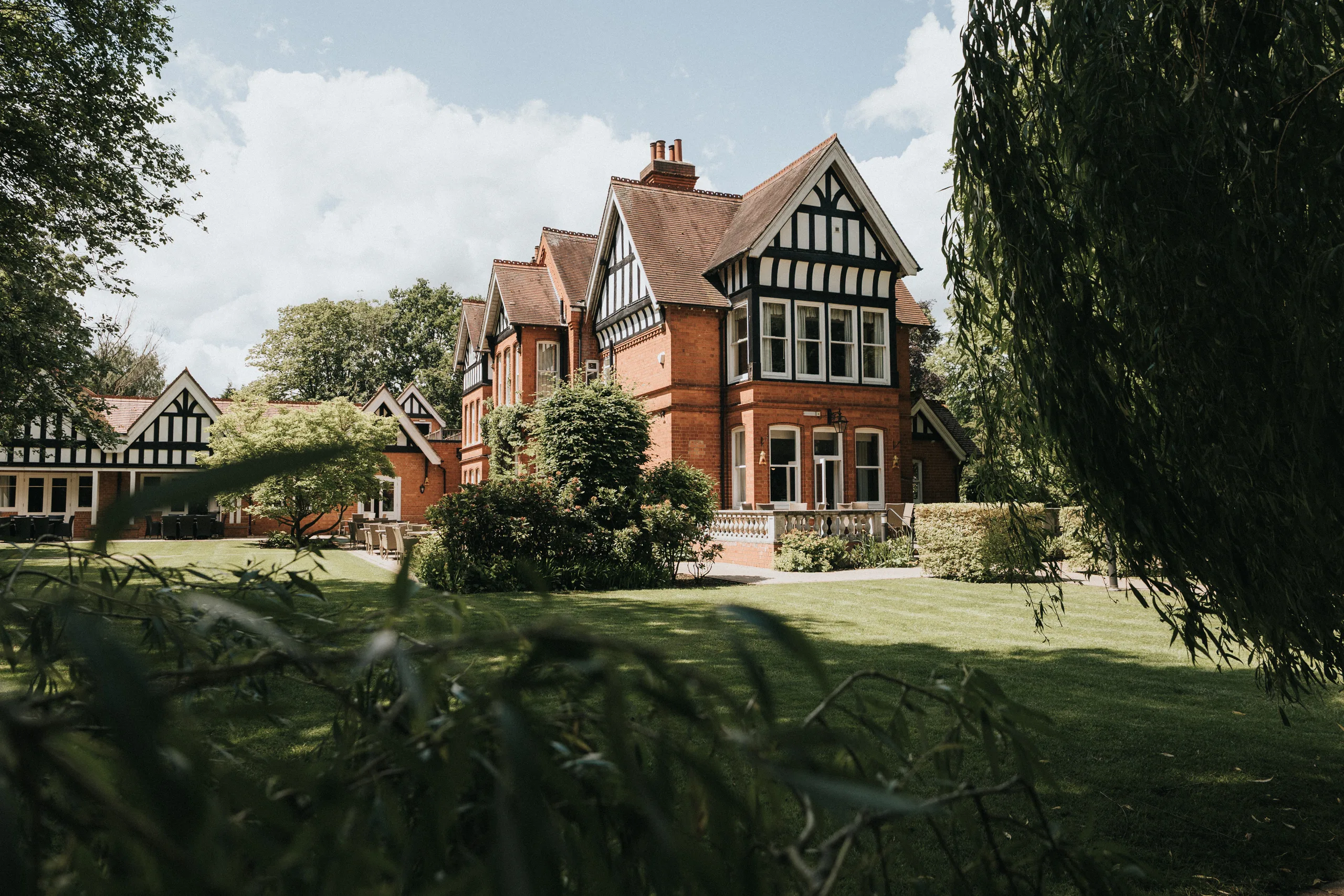 steph-carl-wedding-dower-house-hotel-lincolnshire-woodall-spa-1-of-627-©-aimee-lince-photography-06-06-2025 A large, red-brick Tudor-style house with timber framing sits amid lush green lawns and trees—an idyllic setting for an outdoor wedding in Lincolnshire, perfect for a romantic Woodhall Spa wedding or a wedding at Dower House Hotel. © Aimee Lince Photography - Wedding photographer in Lincolnshire, Yorkshire & Nottinghamshire