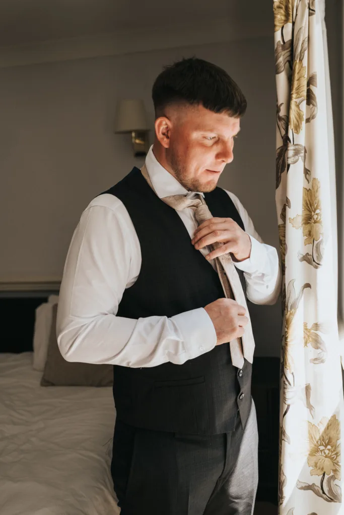 A man in a white shirt and dark vest stands by a window with floral curtains, tying his light-colored tie before a wedding at Dower House Hotel. Sunlight streams in, highlighting his focused expression in the softly lit room. © Aimee Lince Photography - Wedding photographer in Lincolnshire, Yorkshire & Nottinghamshire