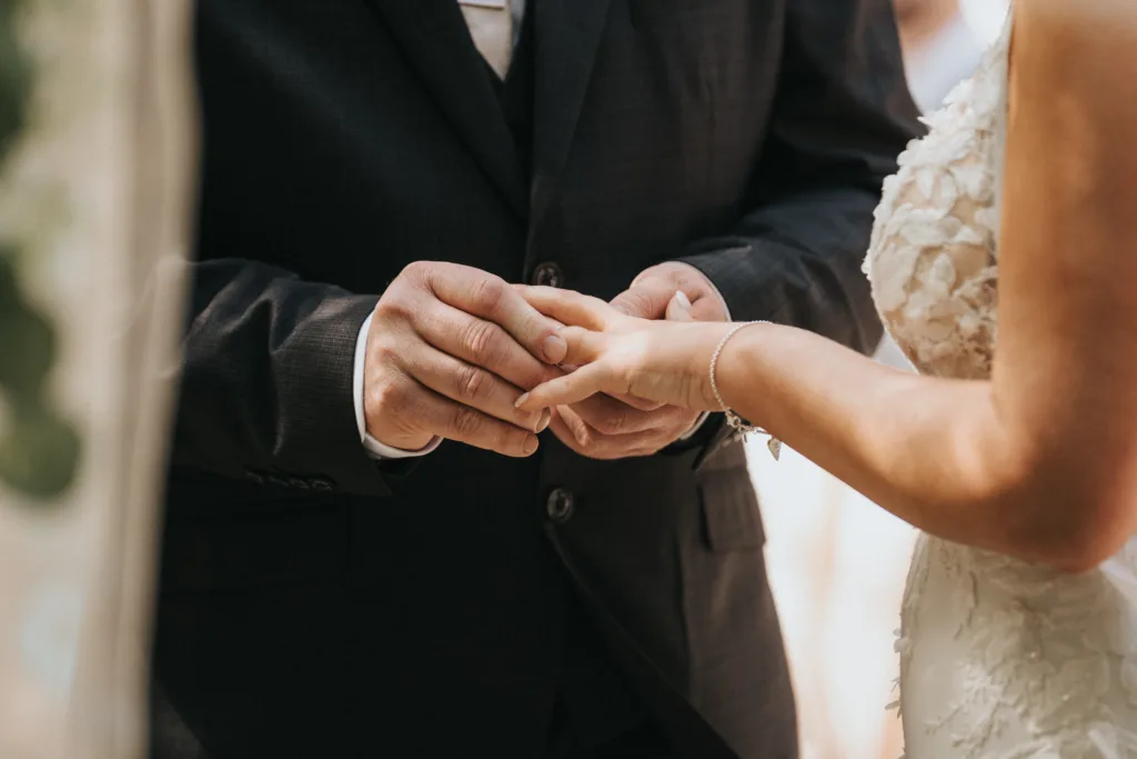 A close-up of a groom in a dark suit gently placing a ring on the bride’s finger during an outdoor wedding Lincolnshire ceremony. The bride wears a textured gown and delicate bracelet, with softly blurred background focusing on their hands. © Aimee Lince Photography - Wedding photographer in Lincolnshire, Yorkshire & Nottinghamshire