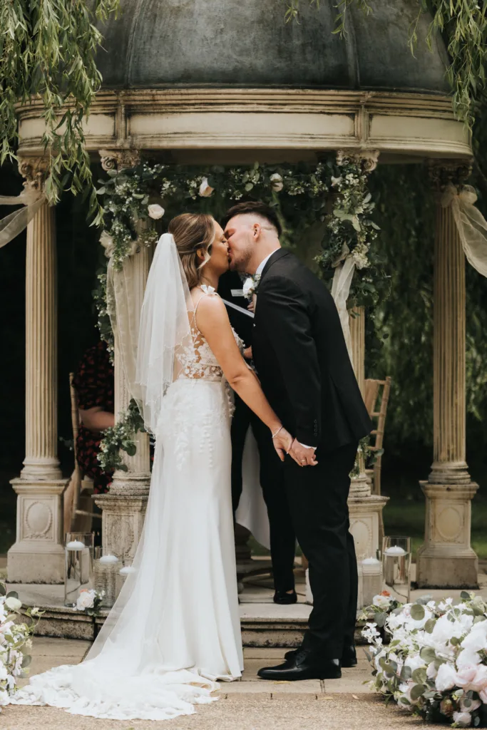 A bride and groom share a kiss under a decorated gazebo during their Dower House Hotel wedding. The bride wears a white dress with a veil, and the groom wears a black suit. They hold hands, surrounded by flowers and greenery. © Aimee Lince Photography - Wedding photographer in Lincolnshire, Yorkshire & Nottinghamshire