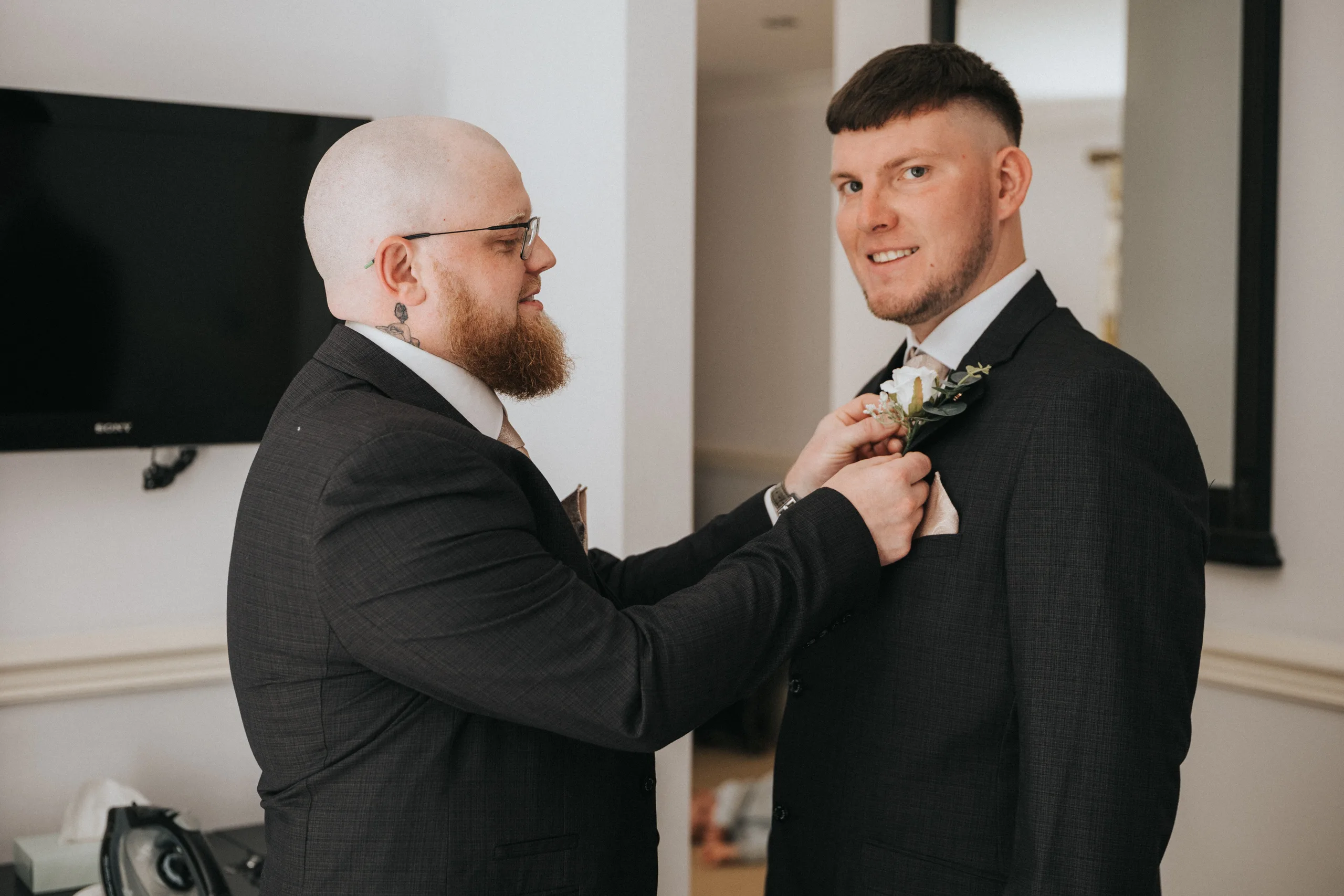steph-carl-wedding-dower-house-hotel-lincolnshire-woodall-spa-15-of-627-©-aimee-lince-photography-06-06-2025 Two men in dark suits are indoors, one adjusting the other’s boutonniere. The man on the left is bald with a beard and glasses, focusing intently. Captured by a Lincolnshire wedding photographer, a TV and mirror are visible in the background. © Aimee Lince Photography - Wedding photographer in Lincolnshire, Yorkshire & Nottinghamshire