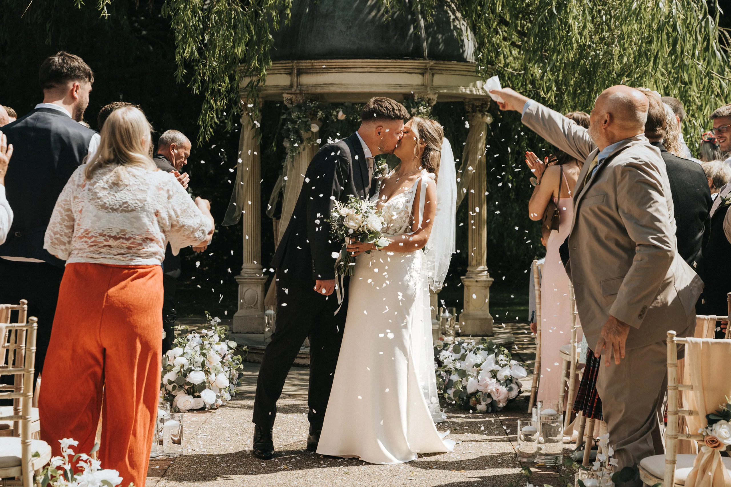 steph-carl-wedding-dower-house-hotel-lincolnshire-woodall-spa-154-of-627-©-aimee-lince-photography-06-06-2025 A bride and groom kiss at a joyful outdoor Dower House Hotel wedding, surrounded by guests tossing white confetti. Bright greenery and flowers set the scene, with chairs lining the aisle for this festive celebration in Woodhall Spa. © Aimee Lince Photography - Wedding photographer in Lincolnshire, Yorkshire & Nottinghamshire