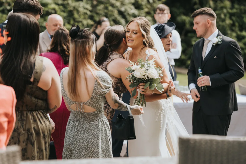 A bride in a white dress and veil, holding a bouquet, smiles while hugging a guest outdoors. Guests in formal attire chat and celebrate as sunlight and greenery surround this joyful Woodhall Spa wedding scene in Lincolnshire. © Aimee Lince Photography - Wedding photographer in Lincolnshire, Yorkshire & Nottinghamshire