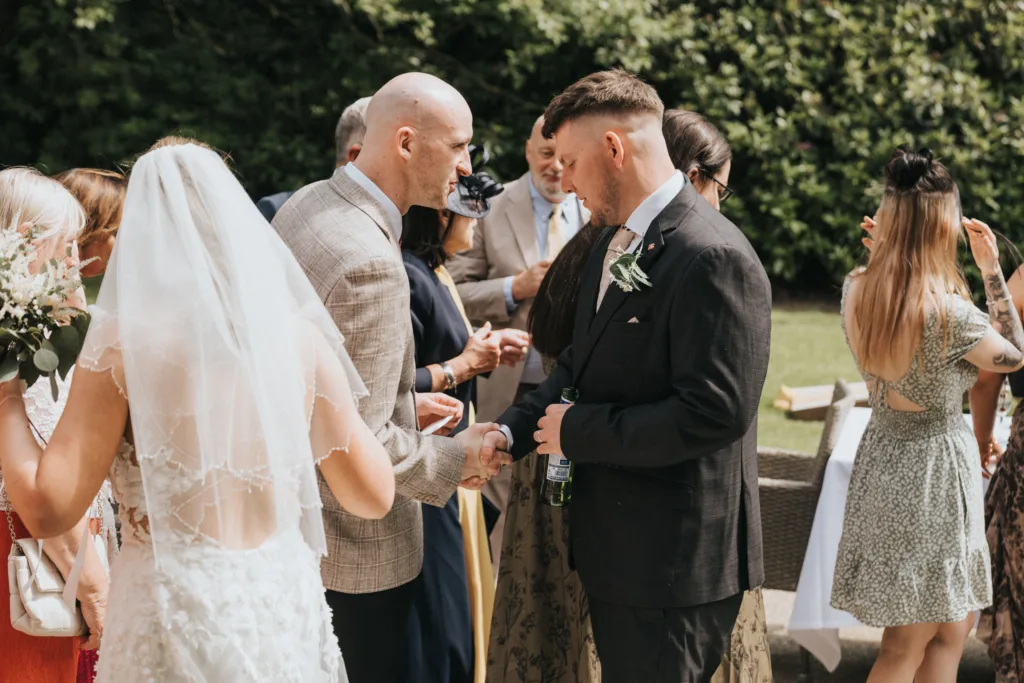 A bride in a white lace dress and veil stands with guests outdoors at a Woodhall Spa wedding. Two men in suits shake hands; one holds a drink. Guests in floral dresses chat nearby, surrounded by green foliage and sunlight in a cheerful, celebratory atmosphere. © Aimee Lince Photography - Wedding photographer in Lincolnshire, Yorkshire & Nottinghamshire
