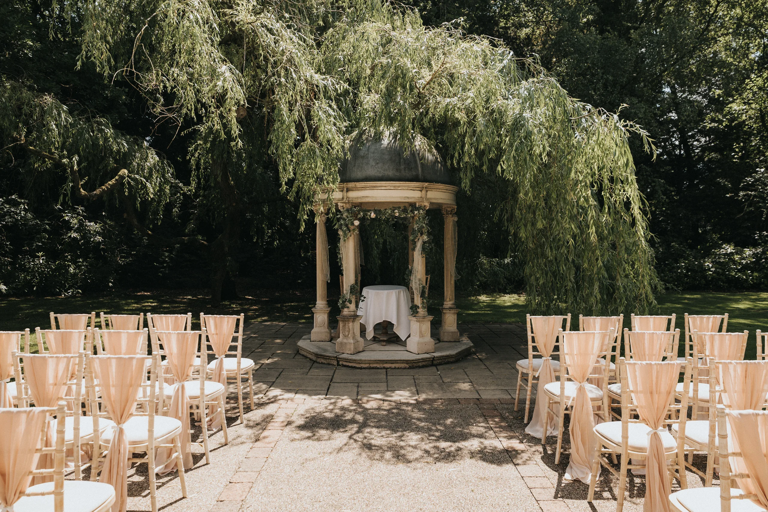 steph-carl-wedding-dower-house-hotel-lincolnshire-woodall-spa-2-of-627-©-aimee-lince-photography-06-06-2025 An outdoor wedding Lincolnshire setup with rows of wooden chairs facing a small stone gazebo draped with greenery. A white-covered table sits inside, surrounded by lush trees and sunlight filtering through the leaves. © Aimee Lince Photography - Wedding photographer in Lincolnshire, Yorkshire & Nottinghamshire