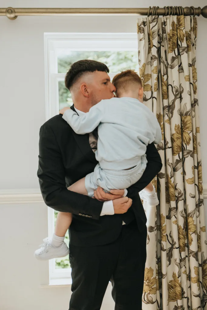 A man in a black suit embraces and kisses a young boy dressed in pale blue near a window with floral curtains, sunlight streaming in—an intimate moment beautifully captured by a Lincolnshire wedding photographer at Dower House Hotel. © Aimee Lince Photography - Wedding photographer in Lincolnshire, Yorkshire & Nottinghamshire