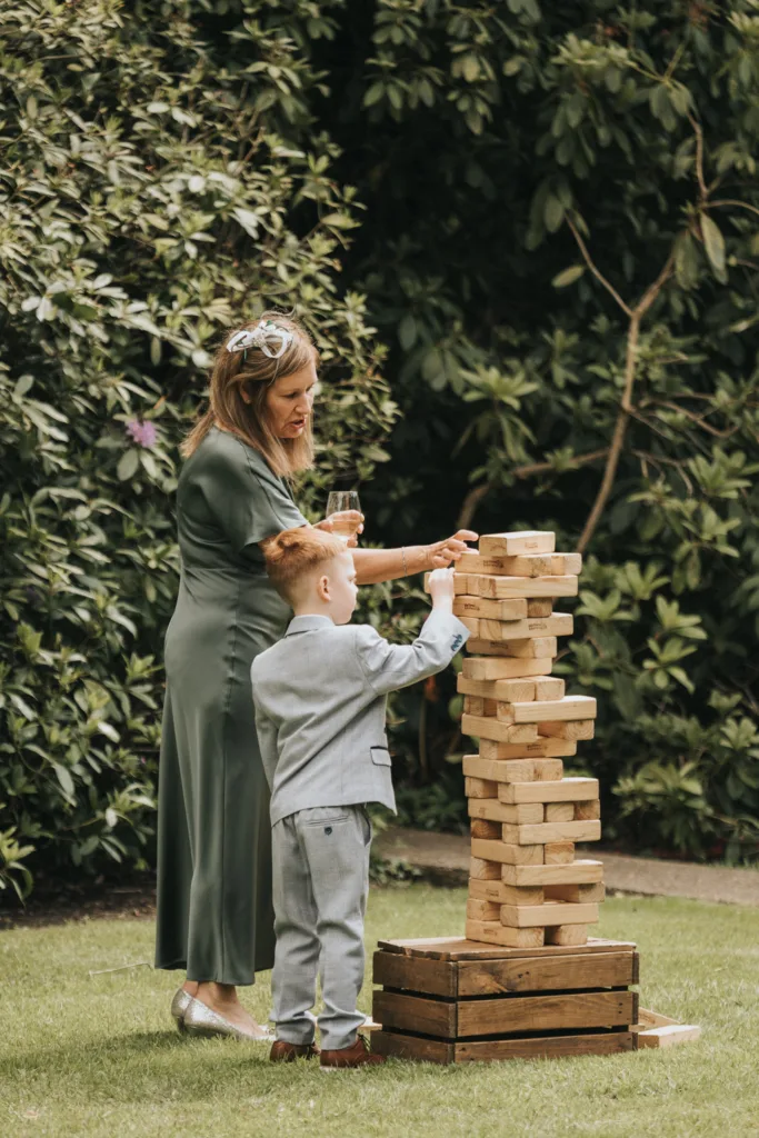 At a Dower House Hotel wedding, a woman in a green dress and a young boy in a grey suit play outdoor Jenga on the grass. She sips her drink, watching as he removes a block, with lush green bushes setting the perfect wedding scene. © Aimee Lince Photography - Wedding photographer in Lincolnshire, Yorkshire & Nottinghamshire