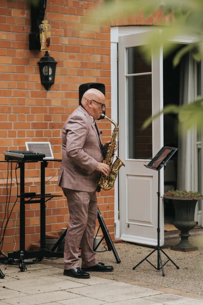 A bald man in a pink plaid suit plays saxophone outside a brick house at a Woodhall Spa wedding. He stands by a keyboard, open door, music stand with sheet music, greenery in the corner, and a black lamp on the wall. © Aimee Lince Photography - Wedding photographer in Lincolnshire, Yorkshire & Nottinghamshire