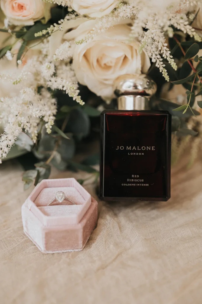 A faceted diamond ring in a pink velvet box sits beside a dark bottle of Jo Malone London Red Hibiscus perfume. Creamy white roses and greenery form the backdrop—perfect for a romantic Woodhall Spa wedding at the Dower House Hotel. © Aimee Lince Photography - Wedding photographer in Lincolnshire, Yorkshire & Nottinghamshire