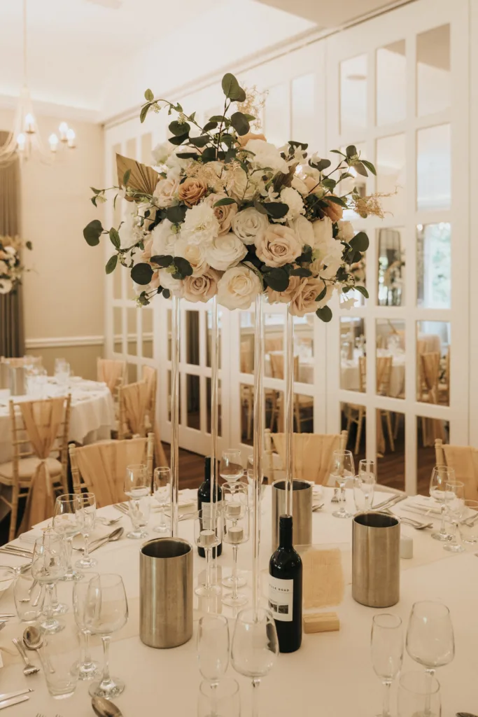 Elegant wedding table setting at a Dower House Hotel wedding, with a round white tablecloth, glassware, wine bottles in metal ice buckets, and a tall floral centerpiece. The bright room features mirrored walls and wooden chairs. © Aimee Lince Photography - Wedding photographer in Lincolnshire, Yorkshire & Nottinghamshire