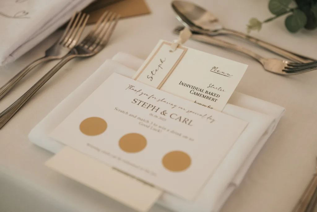 A close-up of a wedding table setting with a fork, spoon, and knife. A folded white napkin holds a personalized card with “STEPH & CARL,” gold scratch-off circles, menu card, and name tag. Perfect for an outdoor wedding Lincolnshire vibe. Greenery in the background. © Aimee Lince Photography - Wedding photographer in Lincolnshire, Yorkshire & Nottinghamshire