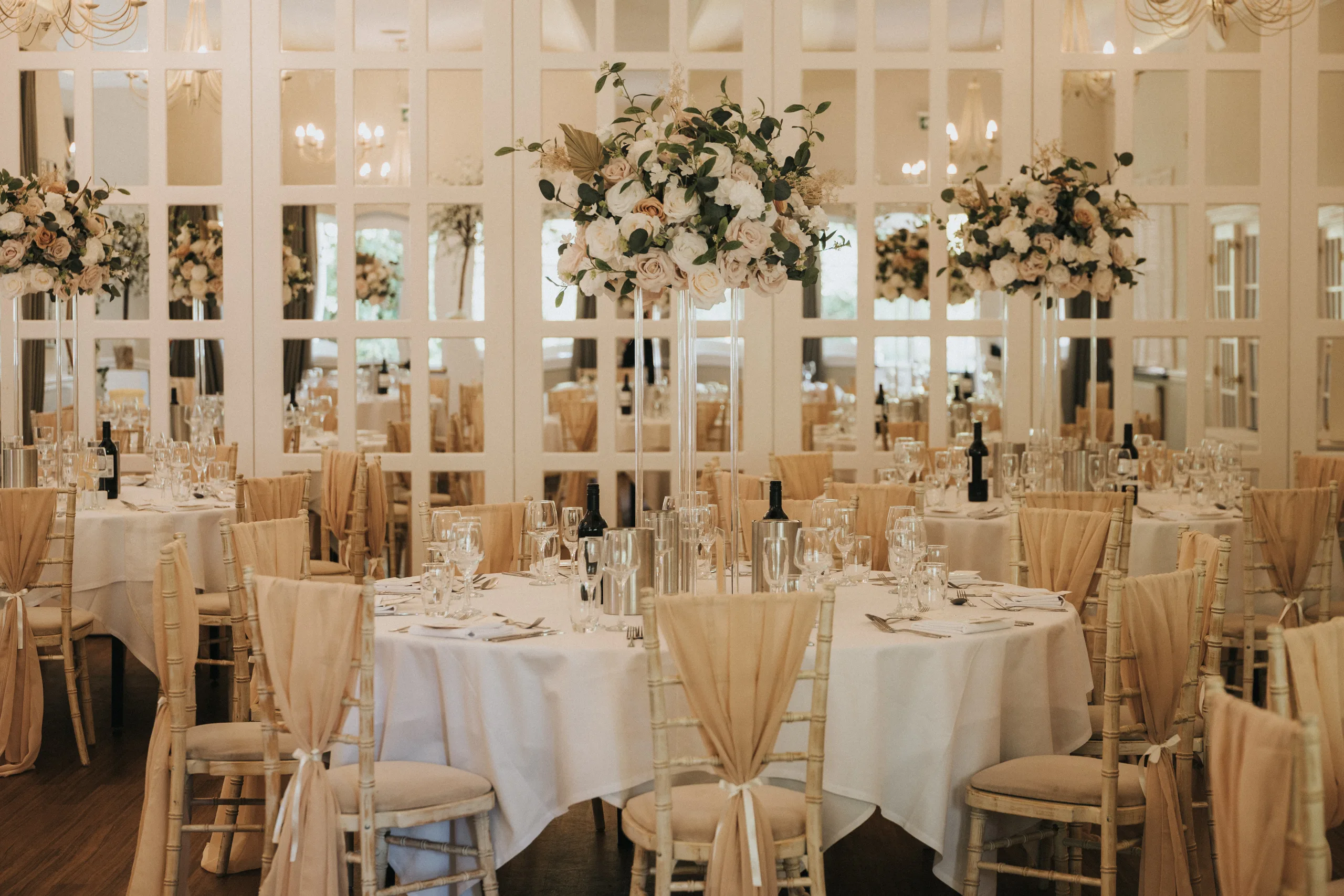 steph-carl-wedding-dower-house-hotel-lincolnshire-woodall-spa-239-of-627-©-aimee-lince-photography-06-06-2025 Elegant Dower House Hotel wedding reception room with round tables draped in white, tall blush and white floral centerpieces, gold chairs, sparkling glassware, and wine bottles; mirrored wall panels reflect the beautiful decor. © Aimee Lince Photography - Wedding photographer in Lincolnshire, Yorkshire & Nottinghamshire