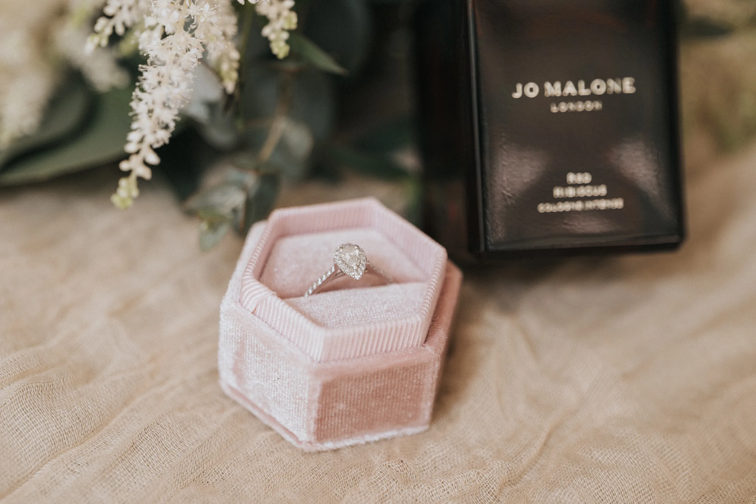 steph-carl-wedding-dower-house-hotel-lincolnshire-woodall-spa-24-of-627-©-aimee-lince-photography-06-06-2025 A pear-shaped diamond engagement ring sits in a soft pink velvet hexagonal box. Nearby, a black Jo Malone London perfume bottle is partially visible, perfect for capturing elegant details at a Dower House Hotel wedding in Woodhall Spa. © Aimee Lince Photography - Wedding photographer in Lincolnshire, Yorkshire & Nottinghamshire