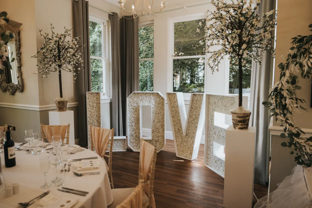 Large white letters spelling "LOVE," adorned with flowers, are displayed by tall windows in an elegant room at a Dower House Hotel wedding. Two flowering trees in pots stand beside them, with a round table set for guests in the foreground. © Aimee Lince Photography - Wedding photographer in Lincolnshire, Yorkshire & Nottinghamshire