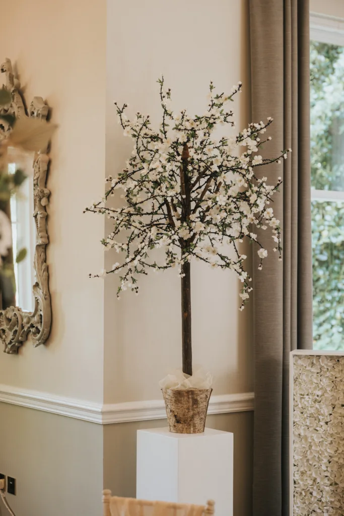 A decorative artificial tree with white blossoms in a rustic pot stands on a white pedestal beside a window with gray curtains—perfect inspiration for a Lincolnshire wedding photographer capturing elegance at a Dower House Hotel wedding. © Aimee Lince Photography - Wedding photographer in Lincolnshire, Yorkshire & Nottinghamshire