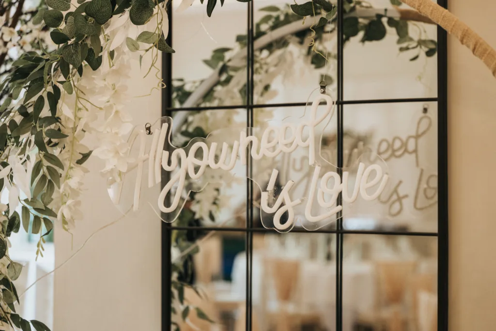 A white script sign reading “All you need is love” hangs in front of a black-framed mirror, surrounded by leafy branches and white flowers—perfect décor inspiration for an outdoor wedding in Lincolnshire, with softly blurred tables and chairs reflected behind. © Aimee Lince Photography - Wedding photographer in Lincolnshire, Yorkshire & Nottinghamshire