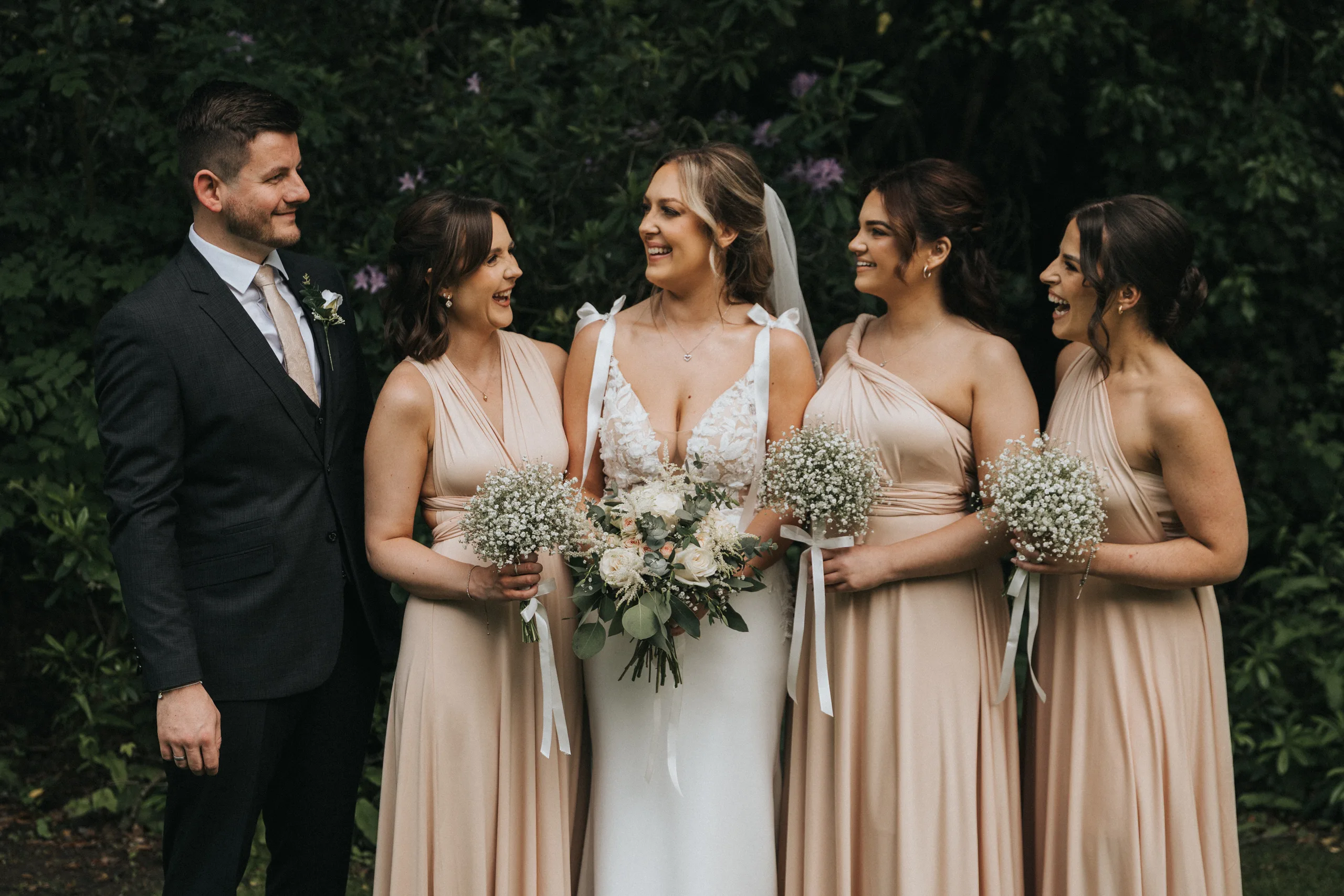 steph-carl-wedding-dower-house-hotel-lincolnshire-woodall-spa-250-of-627-©-aimee-lince-photography-06-06-2025 A bride in a white dress holds a bouquet, standing outside with three bridesmaids in matching blush dresses and a man in a dark suit. All are smiling at each other during an outdoor wedding at the Dower House Hotel, Woodhall Spa. © Aimee Lince Photography - Wedding photographer in Lincolnshire, Yorkshire & Nottinghamshire