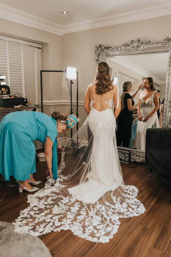 A bride in a white gown with lace train stands facing a large ornate mirror at her Dower House Hotel wedding. Her reflection shows her holding hands with a woman in white, while another in teal adjusts the train. Bright lights illuminate the room. © Aimee Lince Photography - Wedding photographer in Lincolnshire, Yorkshire & Nottinghamshire