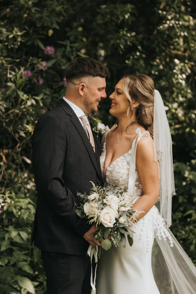 A bride and groom stand close together outdoors, smiling lovingly at each other during their outdoor wedding in Lincolnshire. The bride wears a white dress with floral details, holding a bouquet as green foliage surrounds them in Woodhall Spa. © Aimee Lince Photography - Wedding photographer in Lincolnshire, Yorkshire & Nottinghamshire