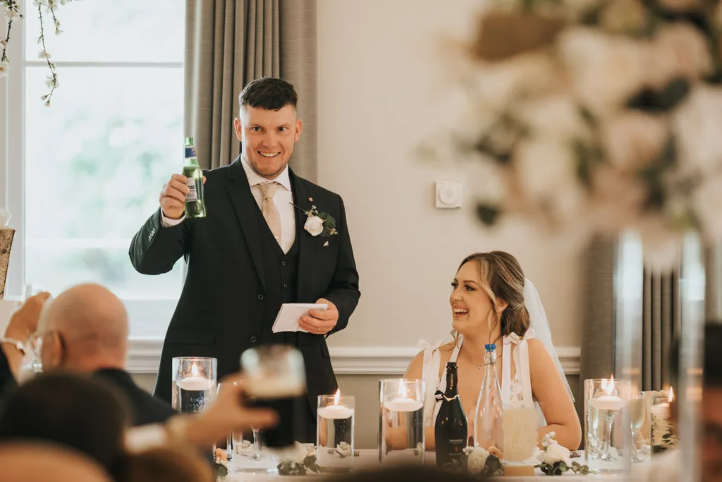 A man in a suit raises a beer bottle in a toast and smiles, while a woman in a wedding dress with a veil sits beside him. At their beautifully decorated table, guests celebrate their Woodhall Spa wedding at Dower House Hotel. © Aimee Lince Photography - Wedding photographer in Lincolnshire, Yorkshire & Nottinghamshire