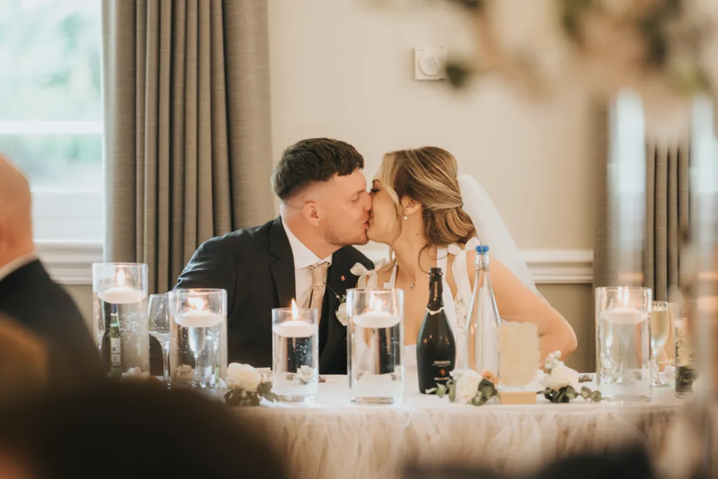 A bride and groom kiss while seated at a Dower House Hotel wedding reception table adorned with white flowers, candles, bottles, and glasses. Both wear formal attire; the bride has a veil. Soft curtains frame the romantic Lincolnshire setting. © Aimee Lince Photography - Wedding photographer in Lincolnshire, Yorkshire & Nottinghamshire