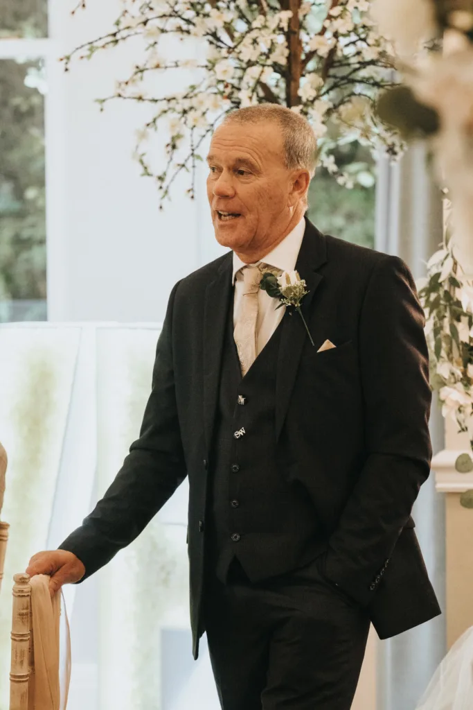 A man in a dark suit with a light tie and boutonnière stands indoors beside a chair, one hand in his pocket. He appears to be speaking at a Dower House Hotel wedding, with decorative trees and soft lighting creating an elegant atmosphere. © Aimee Lince Photography - Wedding photographer in Lincolnshire, Yorkshire & Nottinghamshire