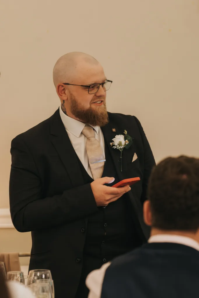 A bearded man in a dark suit, cream tie, and glasses stands indoors at a Dower House Hotel wedding, holding a red phone. He wears a white boutonnière and smiles as if giving a speech—captured perfectly by your Lincolnshire wedding photographer. © Aimee Lince Photography - Wedding photographer in Lincolnshire, Yorkshire & Nottinghamshire