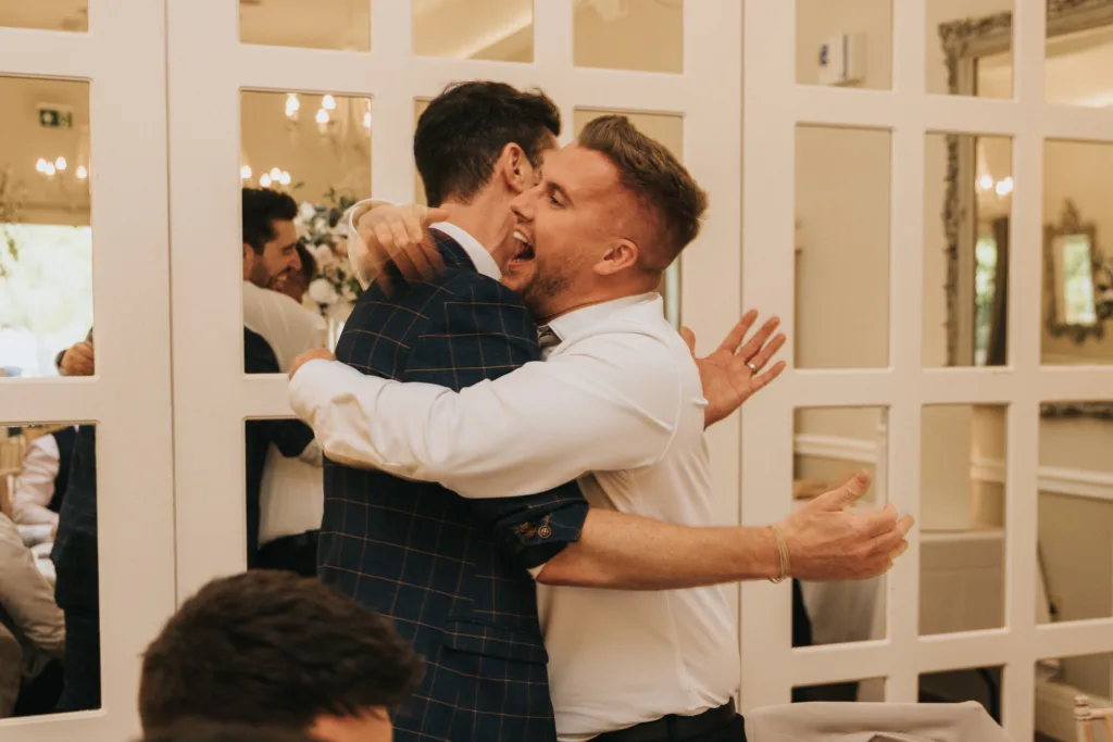 Two men warmly embrace in a bright, elegant room with mirrored wall panels at a Dower House Hotel wedding. One wears a blue plaid suit, the other a white shirt—both joyfully smiling, surrounded by guests and stylish decor. © Aimee Lince Photography - Wedding photographer in Lincolnshire, Yorkshire & Nottinghamshire