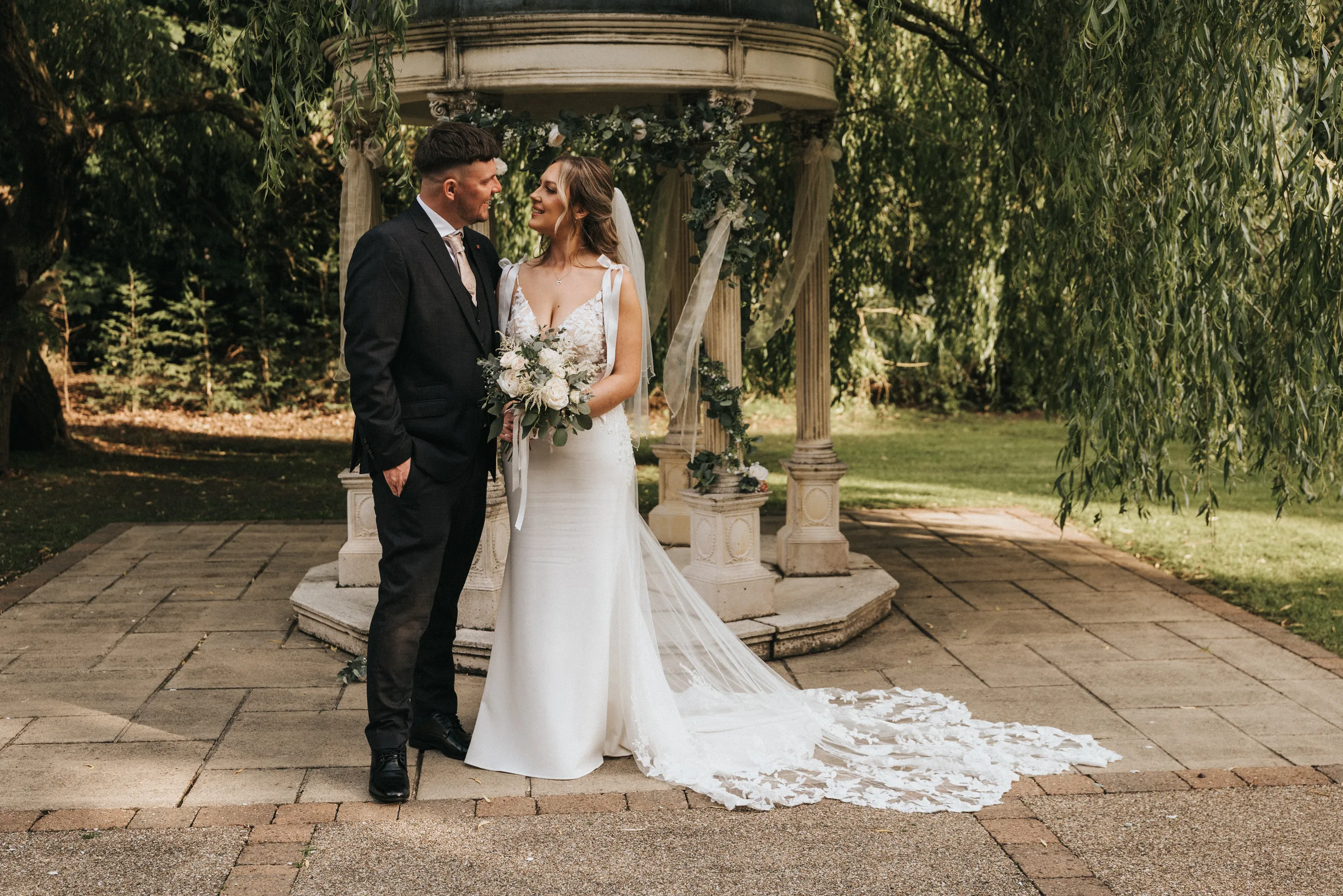 steph-carl-wedding-dower-house-hotel-lincolnshire-woodall-spa-446-of-627-©-aimee-lince-photography-06-06-2025 A bride in a white gown and groom in a black suit stand smiling at each other before a flower-adorned gazebo in a sunlit garden. Captured by a Lincolnshire wedding photographer, her veil trails gracefully behind her at this outdoor wedding in Lincolnshire. © Aimee Lince Photography - Wedding photographer in Lincolnshire, Yorkshire & Nottinghamshire