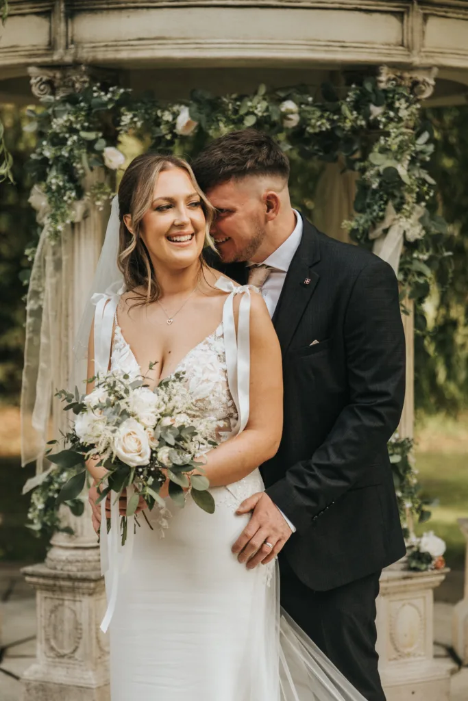 A bride in a white dress holding a bouquet smiles as a groom gently embraces her under a flower-adorned gazebo. Surrounded by greenery, this joyful moment was beautifully captured by a Lincolnshire wedding photographer at an outdoor wedding in Woodhall Spa. © Aimee Lince Photography - Wedding photographer in Lincolnshire, Yorkshire & Nottinghamshire