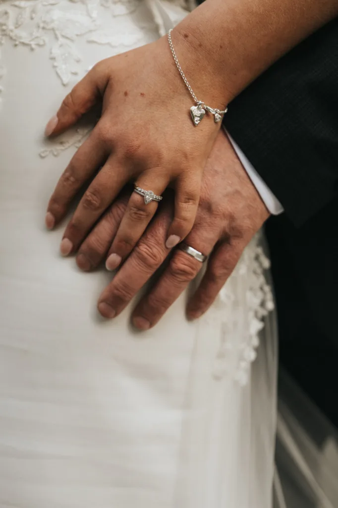 A close-up of two hands resting on a white lace wedding dress at a beautiful Dower House Hotel wedding: a woman's hand with a diamond engagement ring and heart-shaped bracelet gently placed over a man's hand with a simple silver band. © Aimee Lince Photography - Wedding photographer in Lincolnshire, Yorkshire & Nottinghamshire