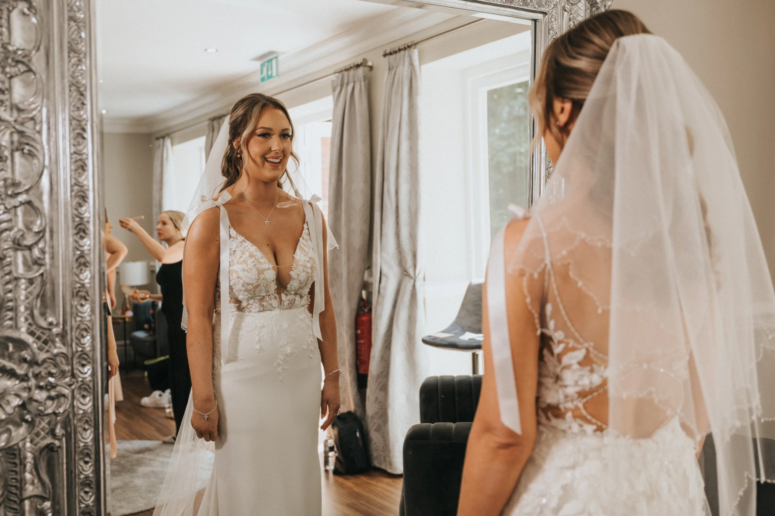 steph-carl-wedding-dower-house-hotel-lincolnshire-woodall-spa-46-of-627-©-aimee-lince-photography-06-06-2025 A bride in a white lace dress and veil smiles at her reflection in a large ornate mirror. The room has light walls, big windows with gray curtains, and women helping her prepare for her Woodhall Spa wedding at the Dower House Hotel. © Aimee Lince Photography - Wedding photographer in Lincolnshire, Yorkshire & Nottinghamshire