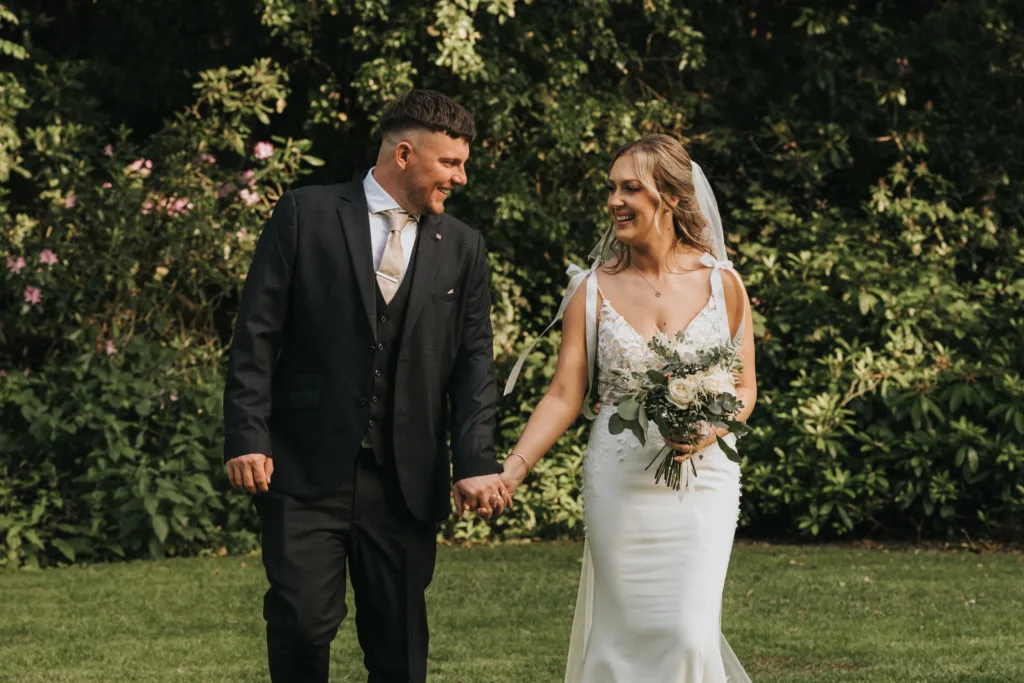 A bride and groom walk hand-in-hand on the grass, smiling at each other during their outdoor wedding in Lincolnshire. The bride holds a bouquet, while lush green foliage hints at a beautiful Dower House Hotel wedding setting. © Aimee Lince Photography - Wedding photographer in Lincolnshire, Yorkshire & Nottinghamshire