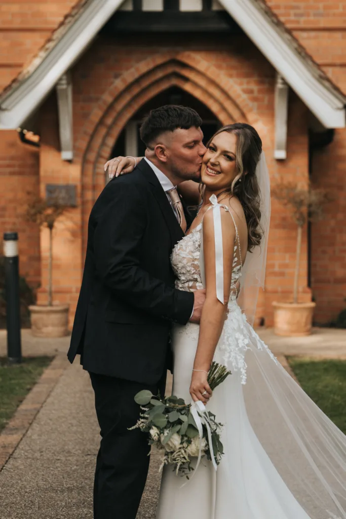 A groom in a black suit kisses his smiling bride on the cheek outside a brick church. The bride, in a white gown with floral details, holds greenery as they stand close, radiating happiness after their Dower House Hotel wedding. © Aimee Lince Photography - Wedding photographer in Lincolnshire, Yorkshire & Nottinghamshire