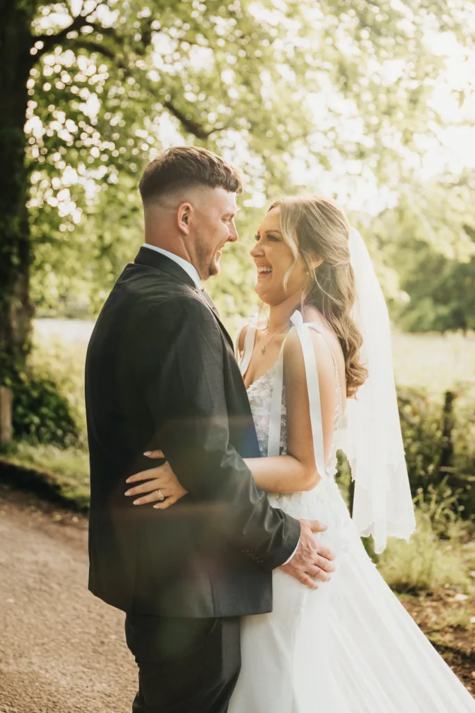 A bride and groom stand outdoors at their Dower House Hotel wedding, smiling lovingly at each other. Sunlight filters through green trees in the background, creating a warm, romantic atmosphere. © Aimee Lince Photography - Wedding photographer in Lincolnshire, Yorkshire & Nottinghamshire