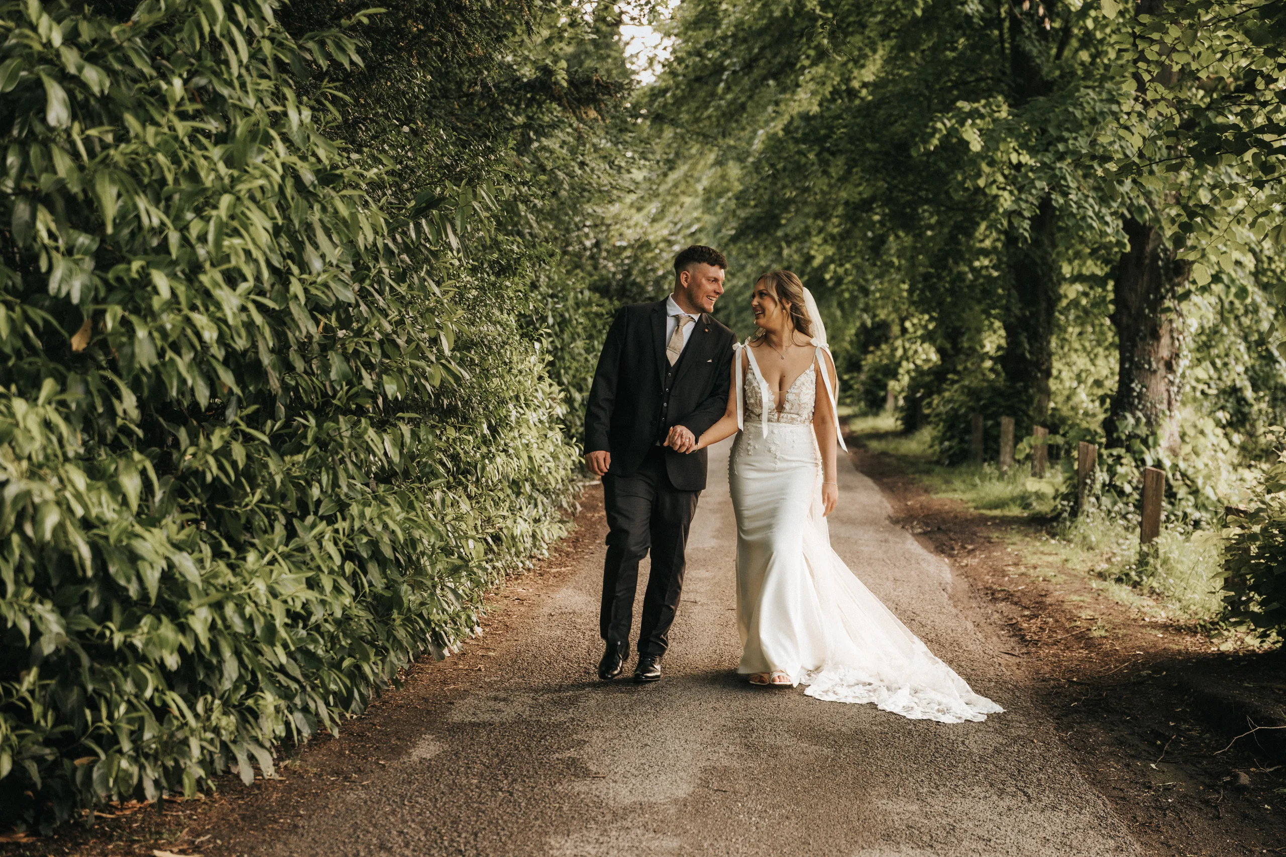 steph-carl-wedding-dower-house-hotel-lincolnshire-woodall-spa-477-of-627-©-aimee-lince-photography-06-06-2025 A bride and groom walk hand in hand down a tree-lined country road after their Woodhall Spa wedding. The groom wears a dark suit and bow tie; the bride wears a white gown with lace details as sunlight filters through the lush green foliage overhead. © Aimee Lince Photography - Wedding photographer in Lincolnshire, Yorkshire & Nottinghamshire