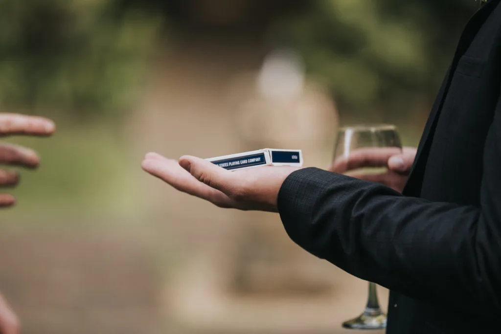 At a Dower House Hotel wedding, a person in a dark suit holds a closed box of playing cards and a wine glass while another hand reaches nearby. The blurred background hints at an elegant outdoor gathering. © Aimee Lince Photography - Wedding photographer in Lincolnshire, Yorkshire & Nottinghamshire
