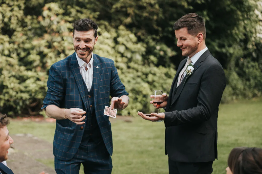 Two men in suits stand outside on grass at a Dower House Hotel wedding. One, smiling, performs a card trick, holding playing cards; the other holds a drink and extends his hand. Blurred guests sit nearby among lush trees and shrubs. © Aimee Lince Photography - Wedding photographer in Lincolnshire, Yorkshire & Nottinghamshire