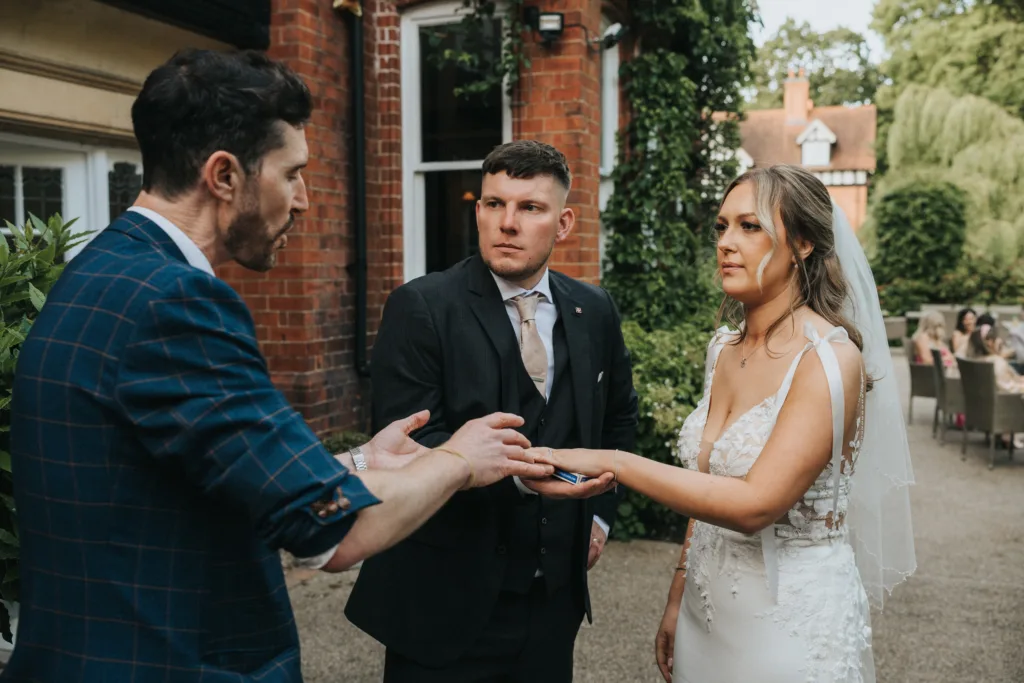 A man in a blue plaid suit performs a trick with a bride and groom during their outdoor wedding in Lincolnshire. The groom, serious in his dark suit, and the bride, in white gown and veil, watch intently near greenery and brick buildings. © Aimee Lince Photography - Wedding photographer in Lincolnshire, Yorkshire & Nottinghamshire