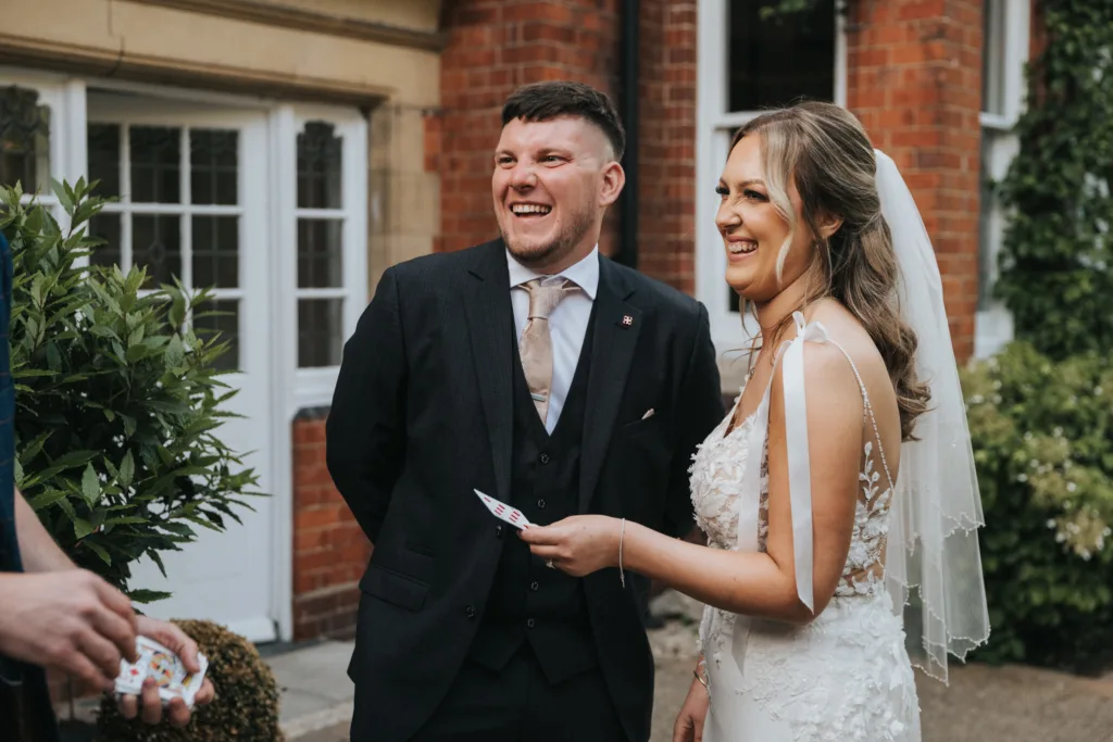 A smiling bride in a white lace dress and veil stands next to her groom, both laughing outdoors near a brick building. The bride holds playing cards, capturing a playful wedding at Dower House Hotel by a Lincolnshire wedding photographer. © Aimee Lince Photography - Wedding photographer in Lincolnshire, Yorkshire & Nottinghamshire