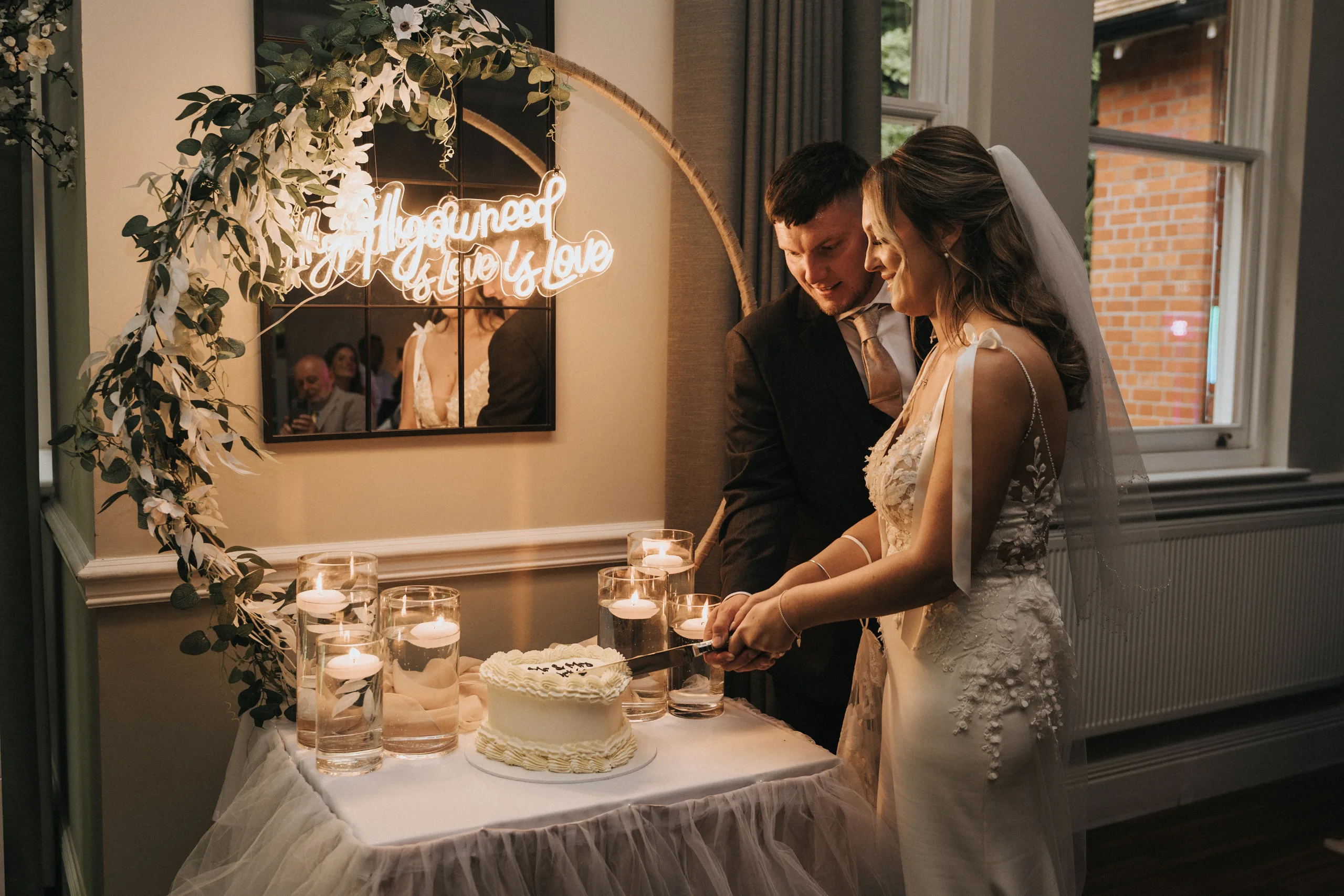 steph-carl-wedding-dower-house-hotel-lincolnshire-woodall-spa-547-of-627-©-aimee-lince-photography-06-06-2025 A bride and groom, both smiling, cut a white wedding cake together on a decorated table with floating candles. Behind them, a neon sign reads “All you need is love.” A leafy arch and guests complete this cozy Dower House Hotel wedding scene. © Aimee Lince Photography - Wedding photographer in Lincolnshire, Yorkshire & Nottinghamshire