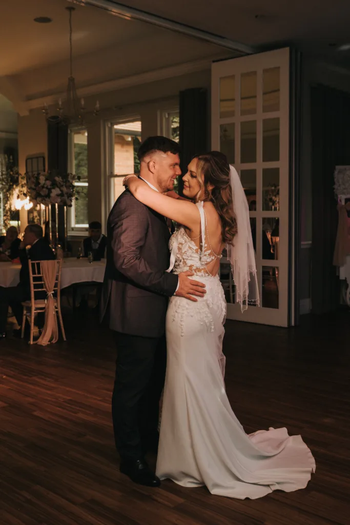 A bride in a white gown and veil and a groom in a suit share their first dance at their Woodhall Spa wedding, embracing on a wooden floor. They smile at each other, surrounded by softly lit tables and floral decorations in the background. © Aimee Lince Photography - Wedding photographer in Lincolnshire, Yorkshire & Nottinghamshire
