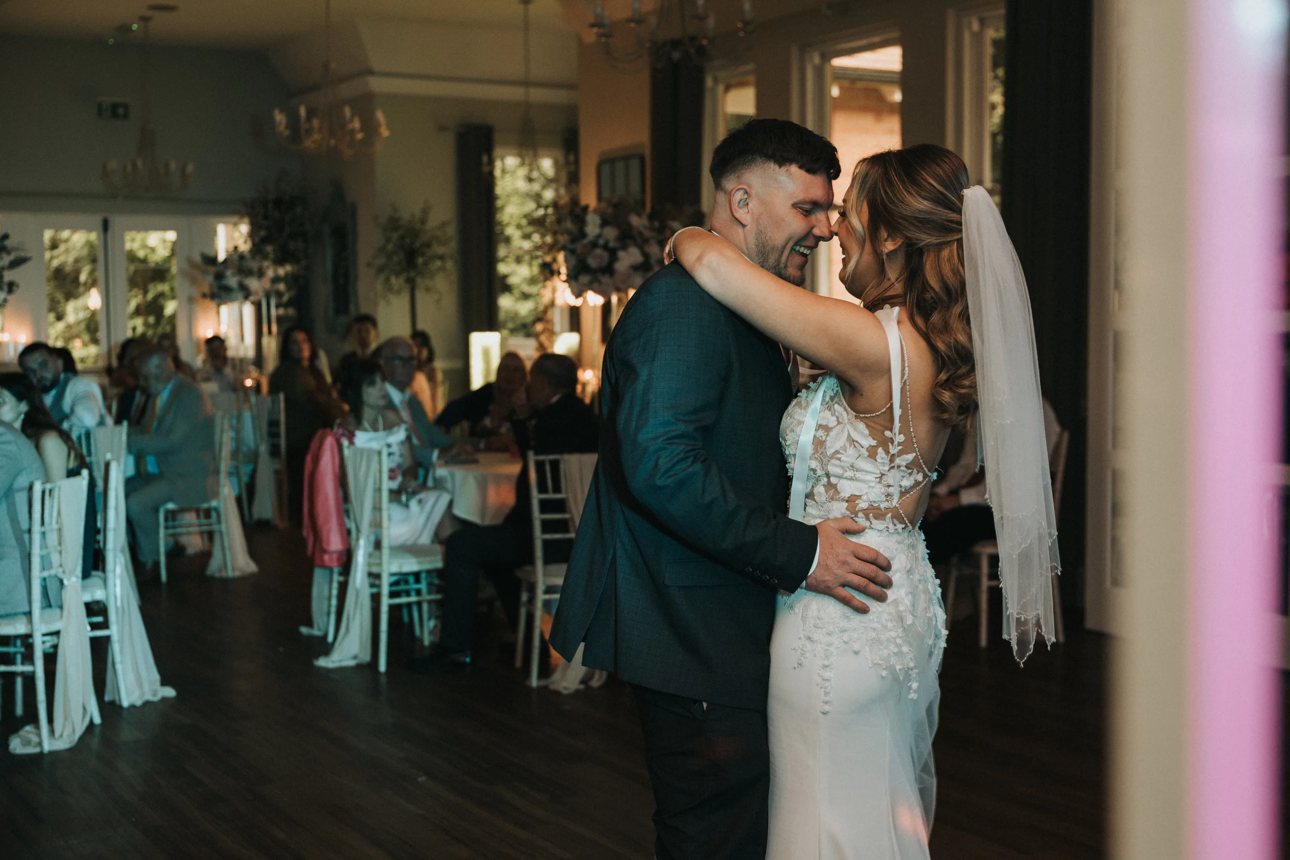 steph-carl-wedding-dower-house-hotel-lincolnshire-woodall-spa-557-of-627-©-aimee-lince-photography-06-06-2025 A bride in a white, lace-back gown and veil shares a joyful first dance with the groom in a dark suit at their elegant Dower House Hotel wedding, surrounded by guests at decorated tables during a warmly lit Woodhall Spa wedding reception. © Aimee Lince Photography - Wedding photographer in Lincolnshire, Yorkshire & Nottinghamshire