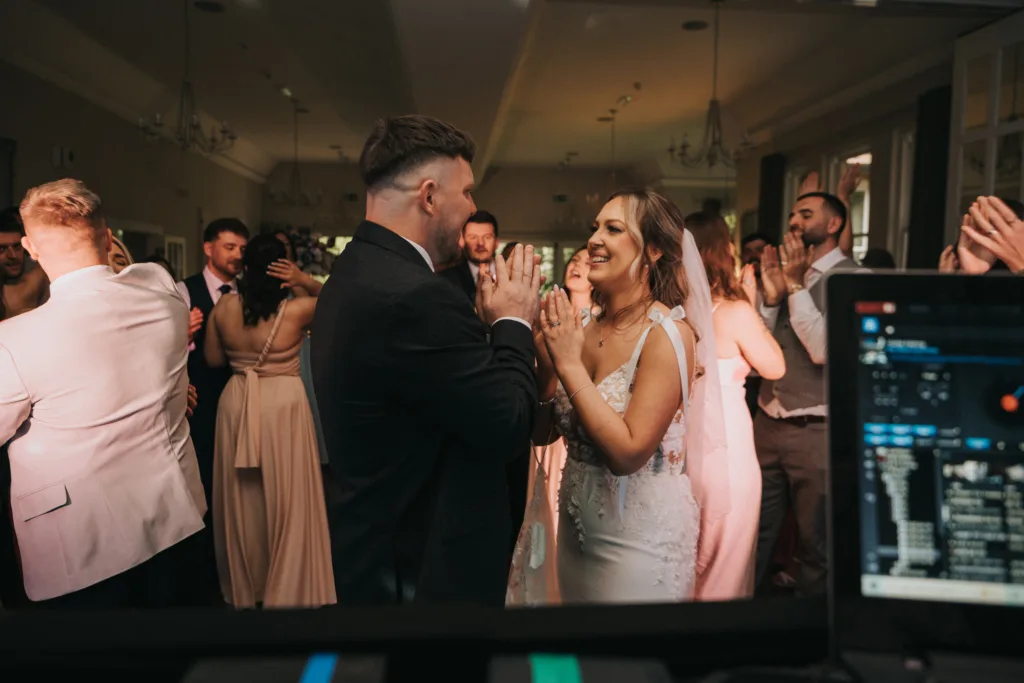 A bride in a white dress and a groom in a dark suit smile and clap hands at their wedding at Dower House Hotel, surrounded by joyful guests dancing. A DJ booth with equipment is in the foreground, adding to the festive ambiance. © Aimee Lince Photography - Wedding photographer in Lincolnshire, Yorkshire & Nottinghamshire