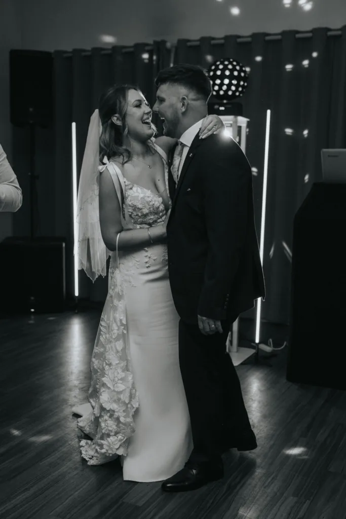A bride in a white dress and veil smiles joyfully while dancing with her groom at their Woodhall Spa wedding. They embrace closely on a wooden floor, surrounded by soft lights—captured by a Lincolnshire wedding photographer in black and white. © Aimee Lince Photography - Wedding photographer in Lincolnshire, Yorkshire & Nottinghamshire