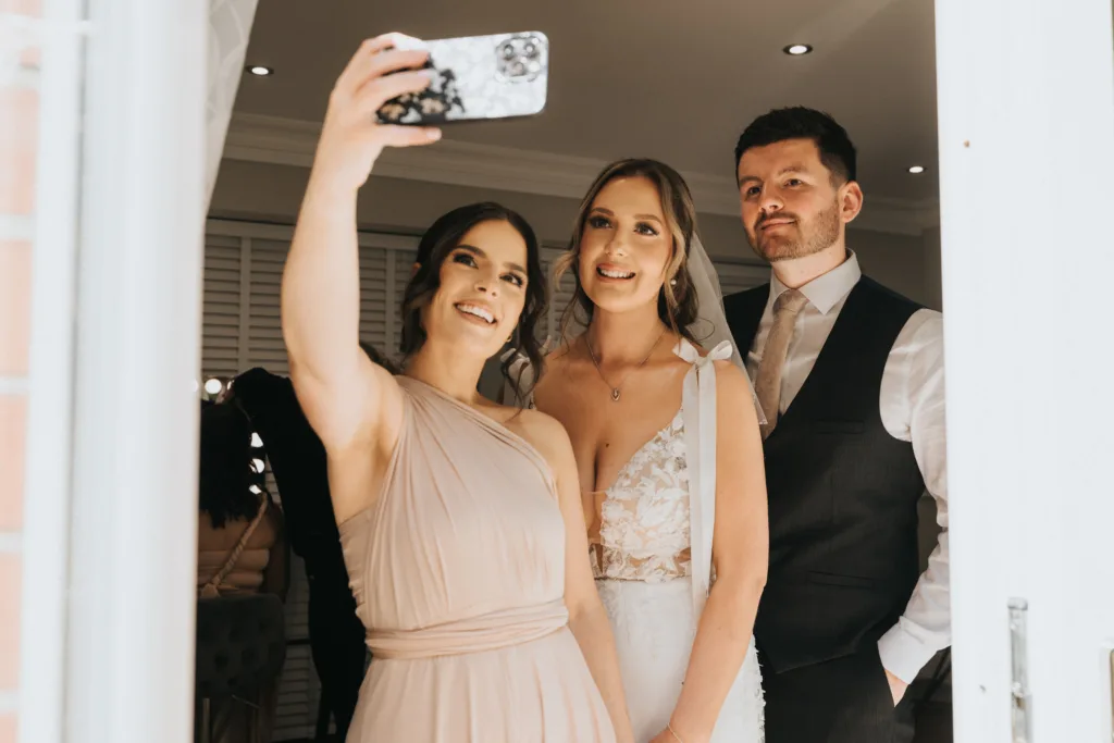 Three people pose for a selfie indoors: a smiling woman in light pink holds the phone, beside a bride in a lace wedding gown and a man in a dark suit. They celebrate their wedding at Dower House Hotel, framed by soft lighting and joy. © Aimee Lince Photography - Wedding photographer in Lincolnshire, Yorkshire & Nottinghamshire